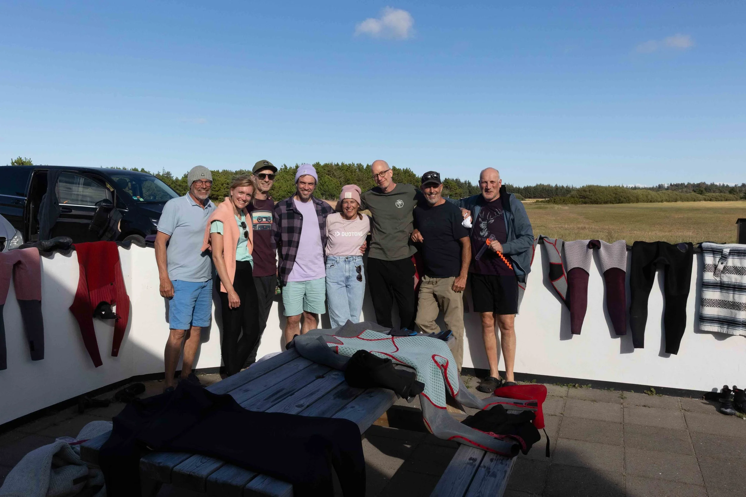Gruppe von neun Menschen, die zusammen auf einer Terrasse stehen, mit Kleidung und Ausrüstung für eine Outdoor-Aktivität, im Hintergrund ein offenes Feld und blauer Himmel.