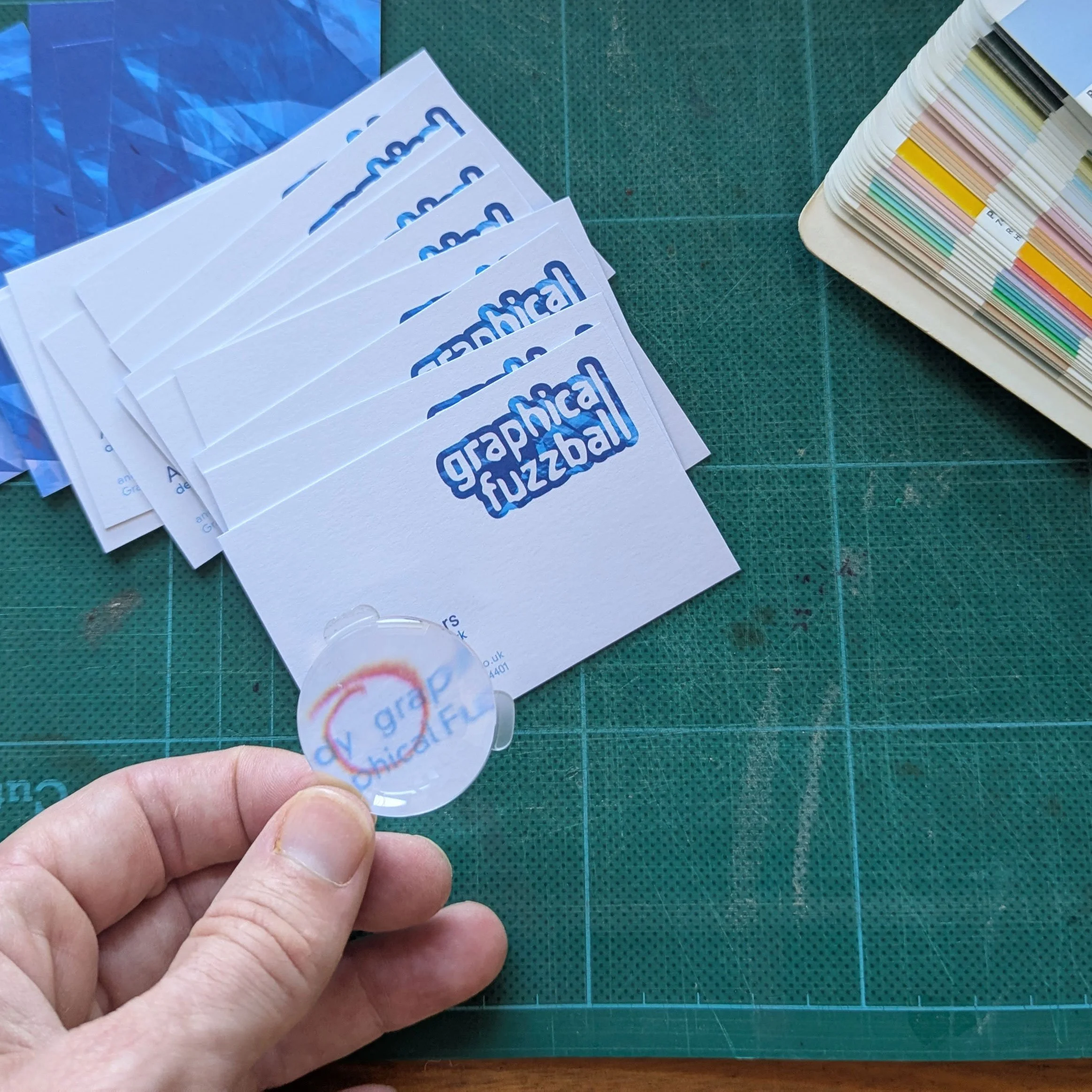 A hand holds a loupe magnifier over some business cards. There is a red pencil circle over a missing @ symbol. The card are sitting on a green cutting mat and there is a swatch book to the right.