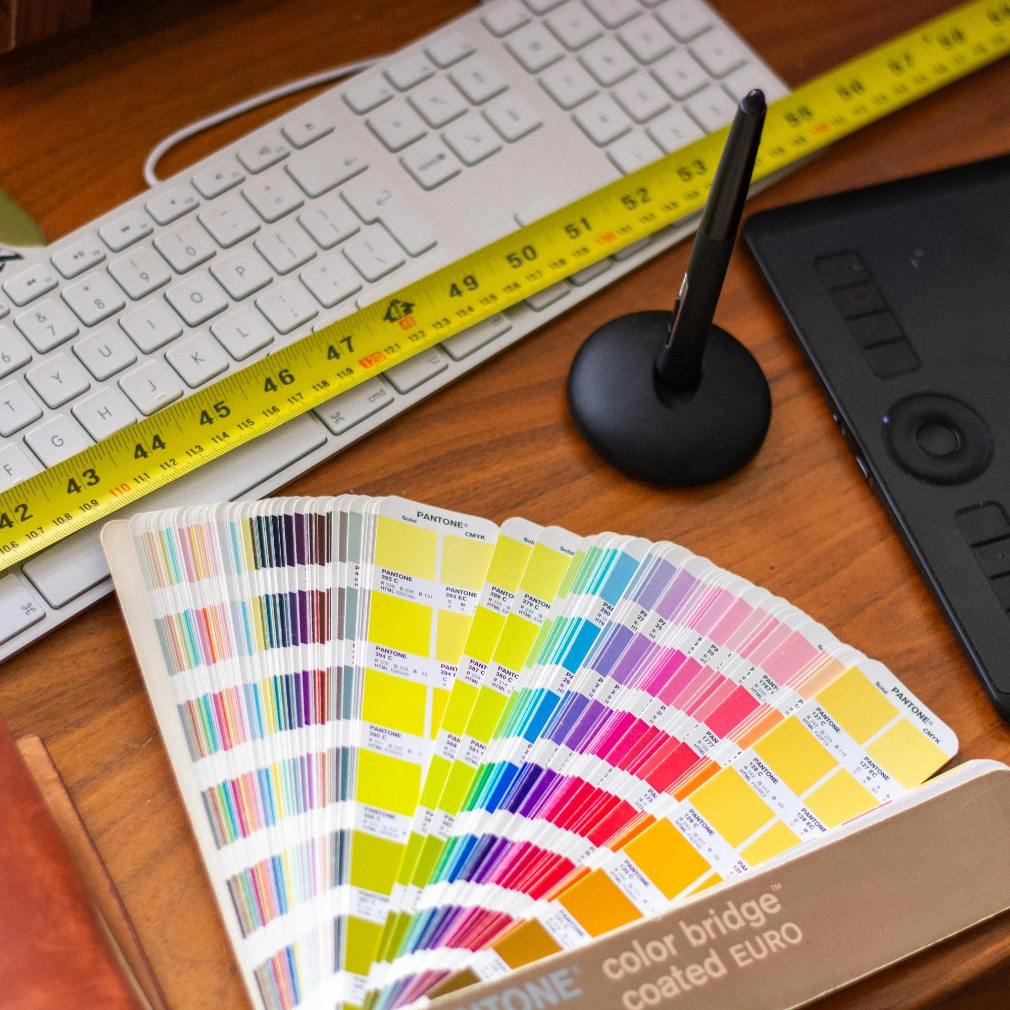 Still life with fanned out pantone swatch book, wacom tablet and pen, apple keyboard and a yellow tape measure sitting on a mid century modern varnished wood desktop.