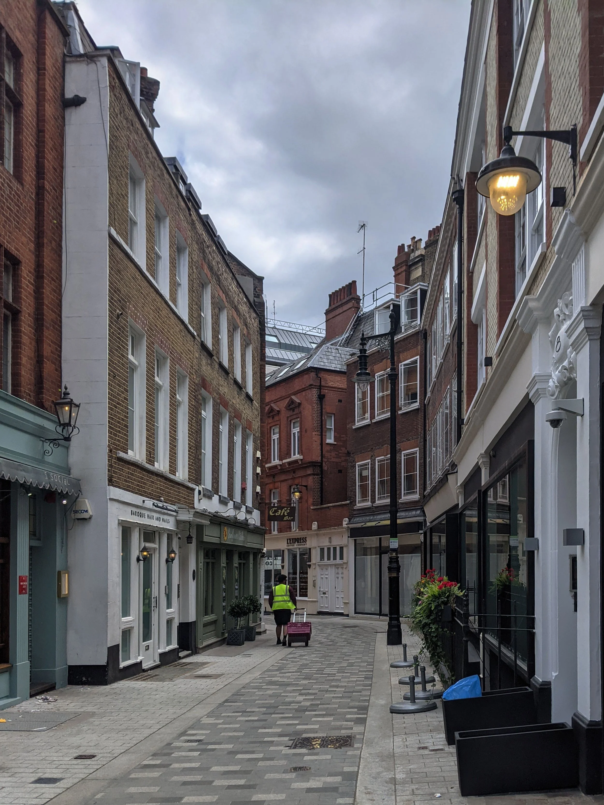 A view down a narrow London side street with a variety of old townhouses and shop fronts. The sky is overcast and a single old style street lamp is glowing as a delivery person pulls a cart around the corner at the end.