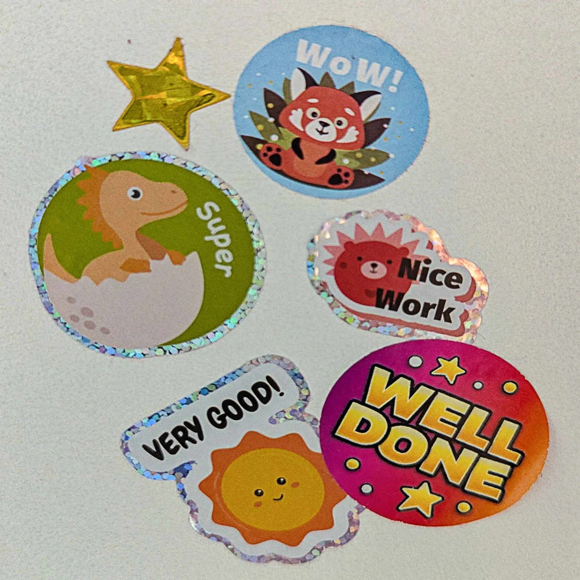 A bunch of stickers on my son's wall. There's a gold star, a red panda that says wow, a dino in an egg shell that says super, a big one that says well done and a couple more brightly coloured ones with similar messages.