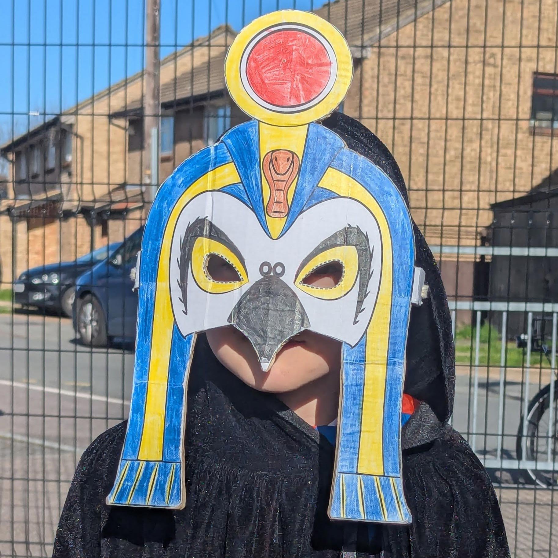 My young lad standing in front of the school fence with houses in the background. It is Egyptian day so he's wearing a black cape over his uniform and a home made mask of the sun god Ra.