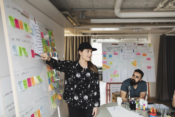 Woman presenting in front of a whiteboard filled with colored post-it notes while two people listen during a meeting.
