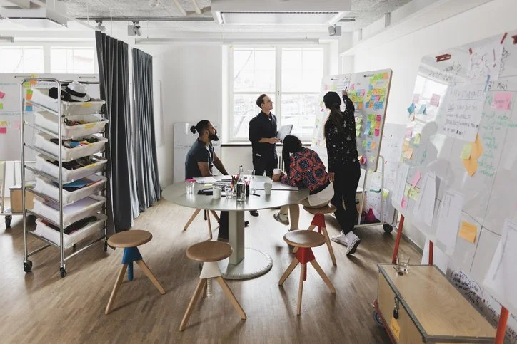 A group of people are working together in a meeting room with whiteboards covered in colorful notes and sticky notes.