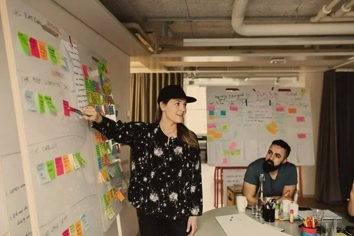 En kvinde gestikulerer mod en tavle med farverige post-its, mens en mand lytter opmærksomt. Begge er i et mødelokale.