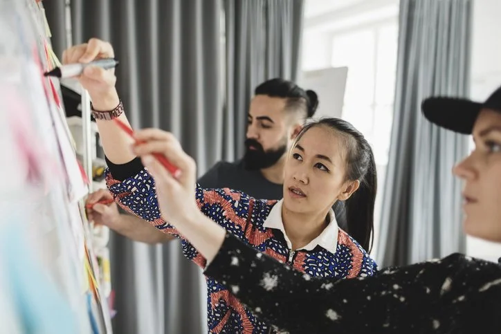 Three people are working on a wall covered with Post-it notes, focusing on a woman writing on a Post-it note.