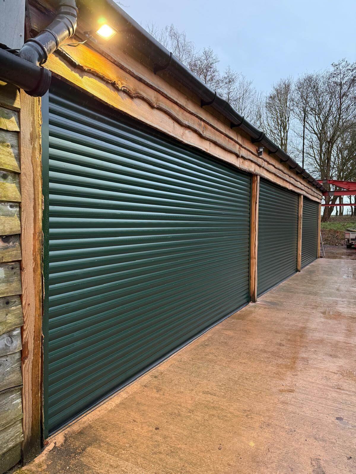 A building with three green metal garage doors, wooden framing, a black roof gutter, and an outdoor light on, with an overcast sky and leafless trees in the background.