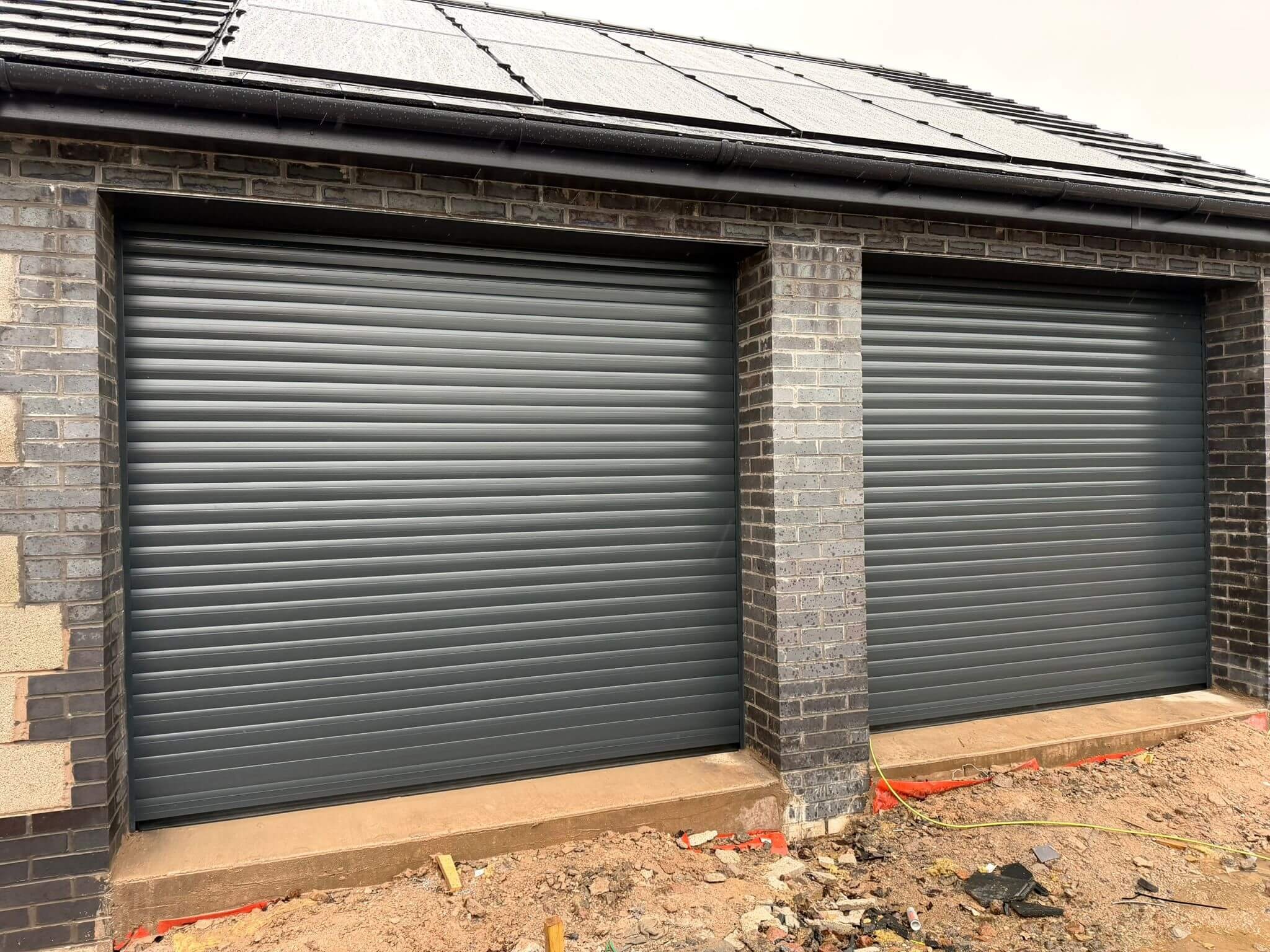 Two closed gray metal garage doors on a house with brick exterior, construction debris and hoses on the ground in front.