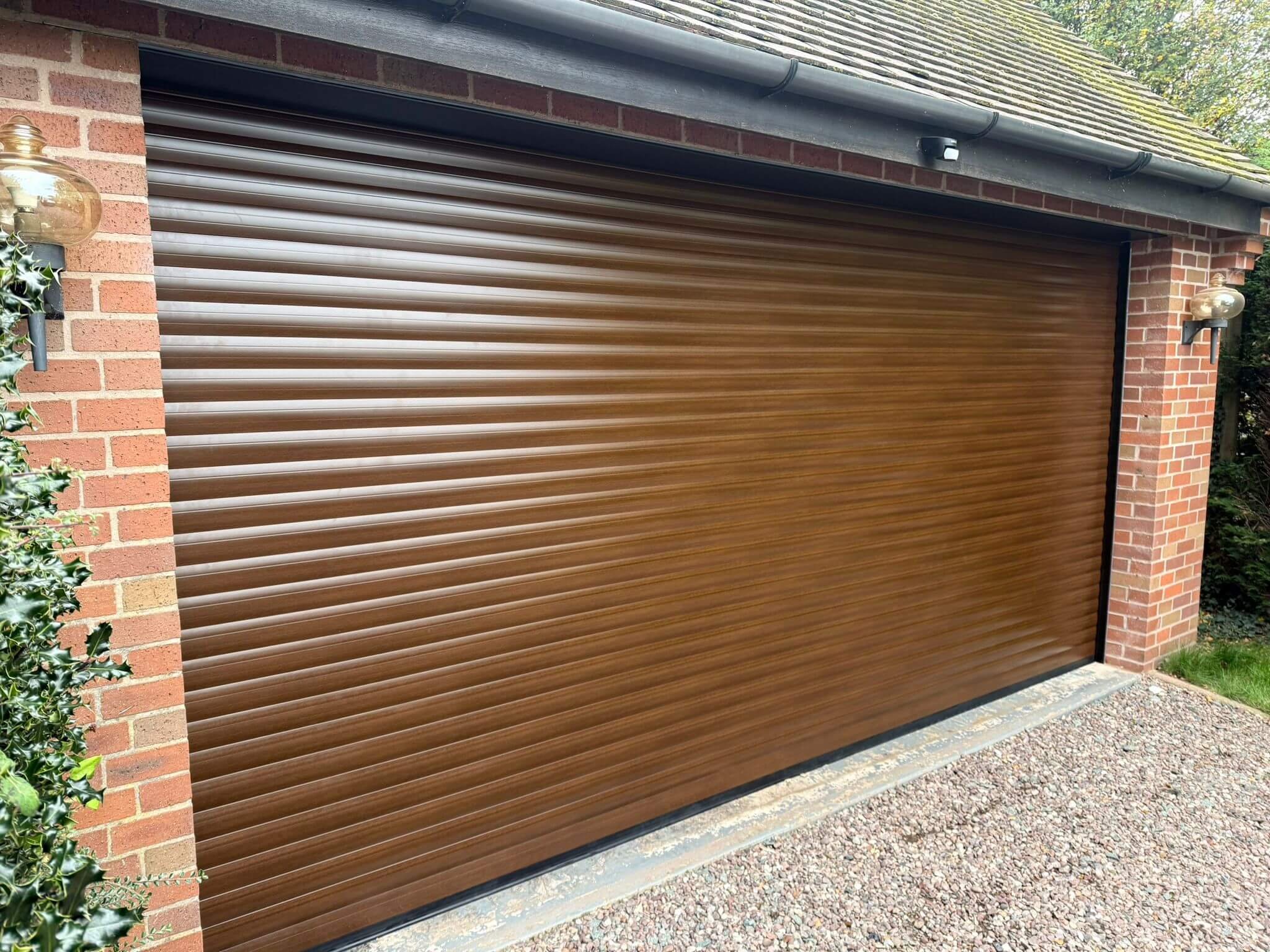 A closed brown roll-up garage door on a brick house with outdoor wall lights and a gravel driveway.