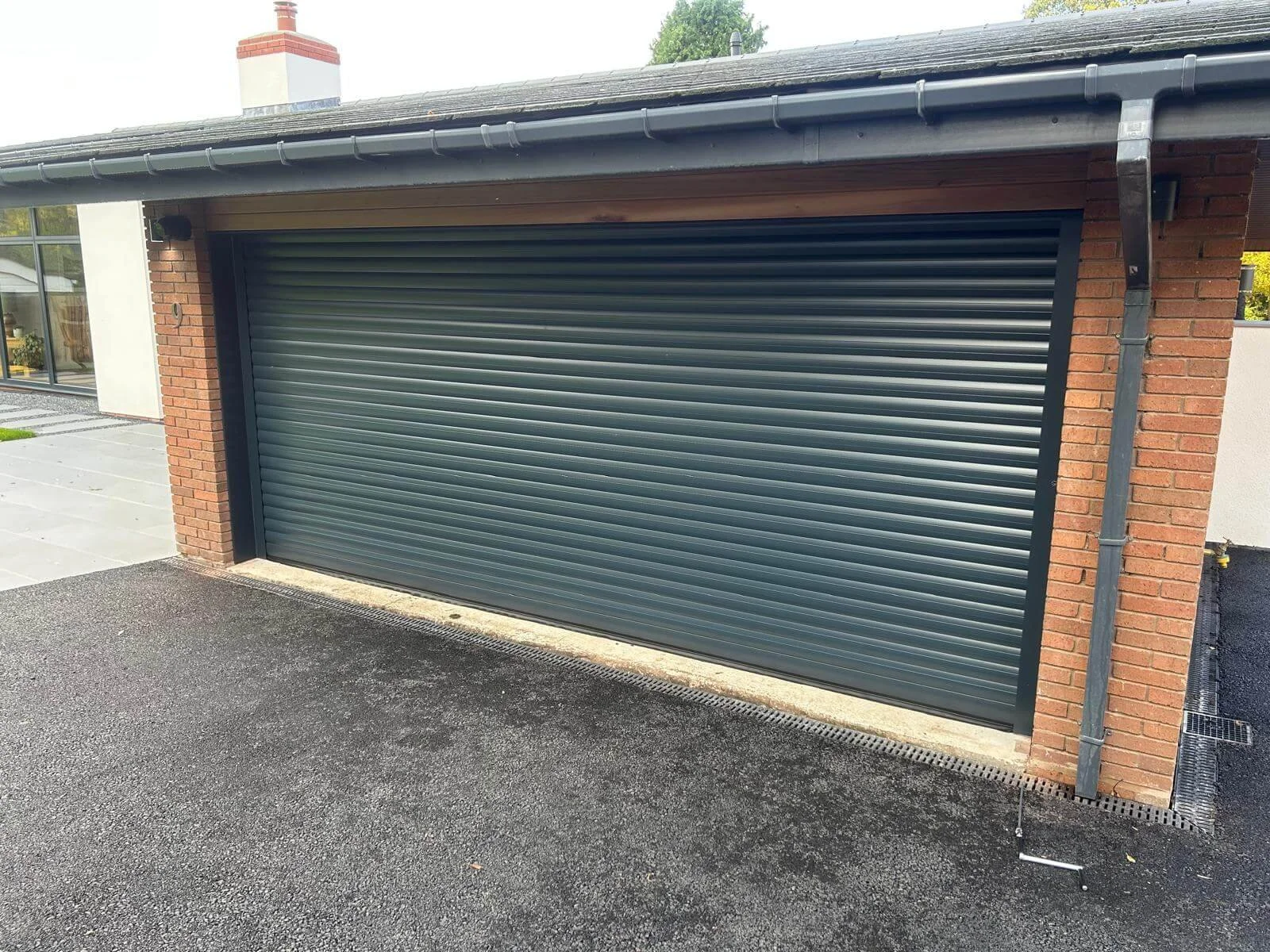 A modern brick garage with black rolling shutter door, located beside a paved driveway and a house with large window. The garage has a metal gutter and downspout.