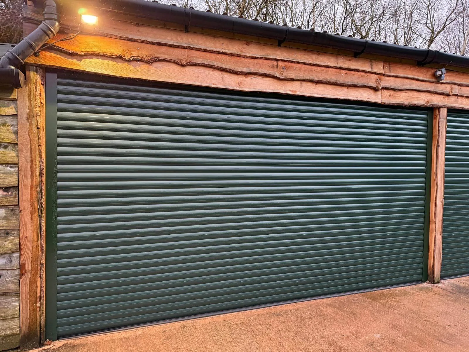 Green metal roll-up garage door with wooden framing and a black roof, outside a building