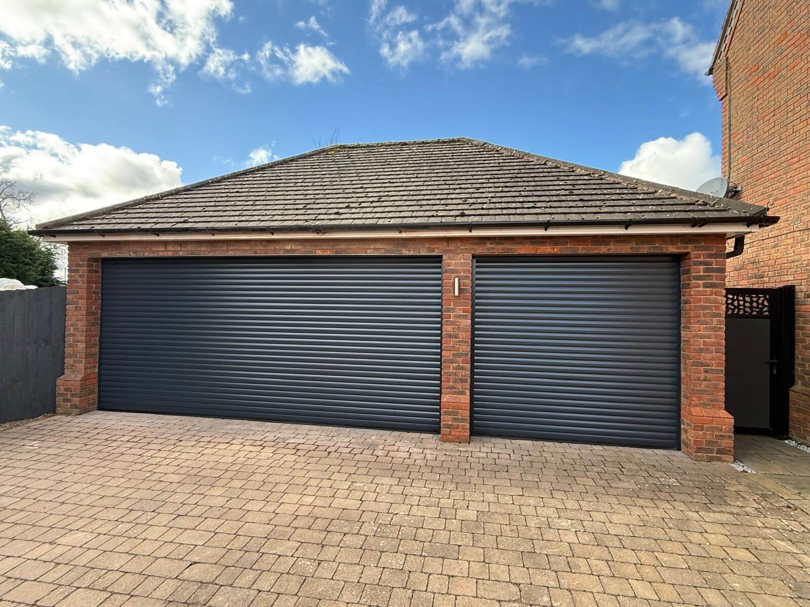 A brick garage with two closed dark gray roller doors, a paved driveway, and a small black gate on the right side, under a blue sky with some clouds.