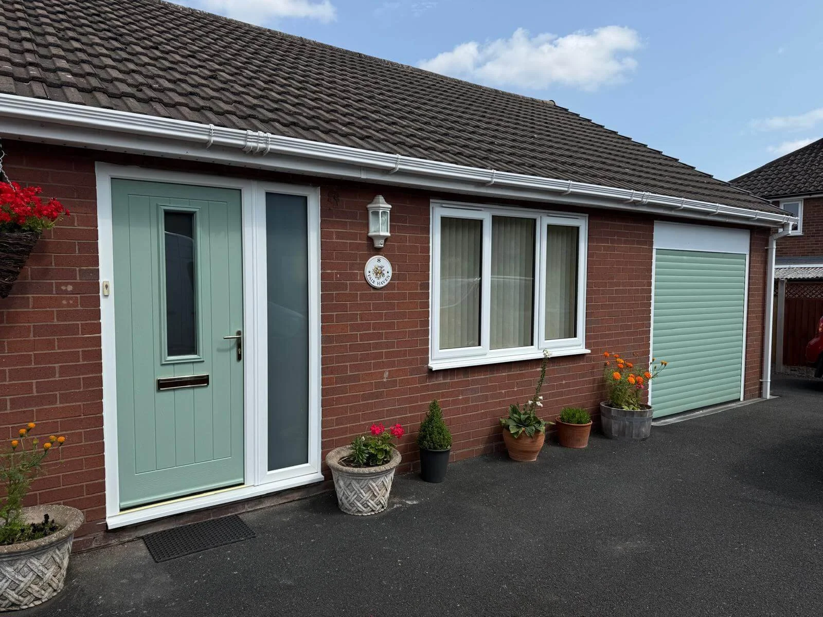 A brick house with a green front door and a large window with blinds. There are potted plants and flowers outside, and a garage with a green roll-up door. The house has a sloped roof with tiles, and a white gutter system.
