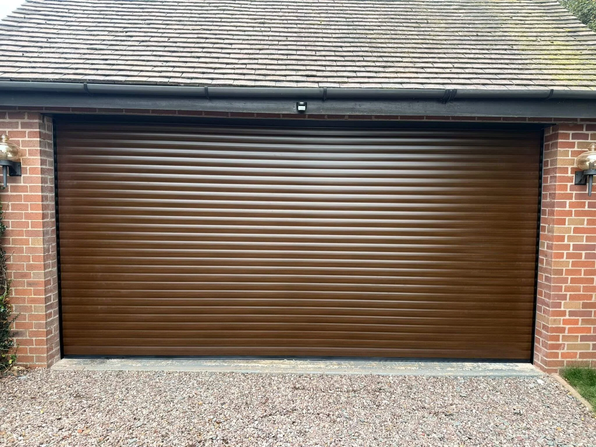Brown metal garage door with brick walls on either side and a gravel driveway in front.