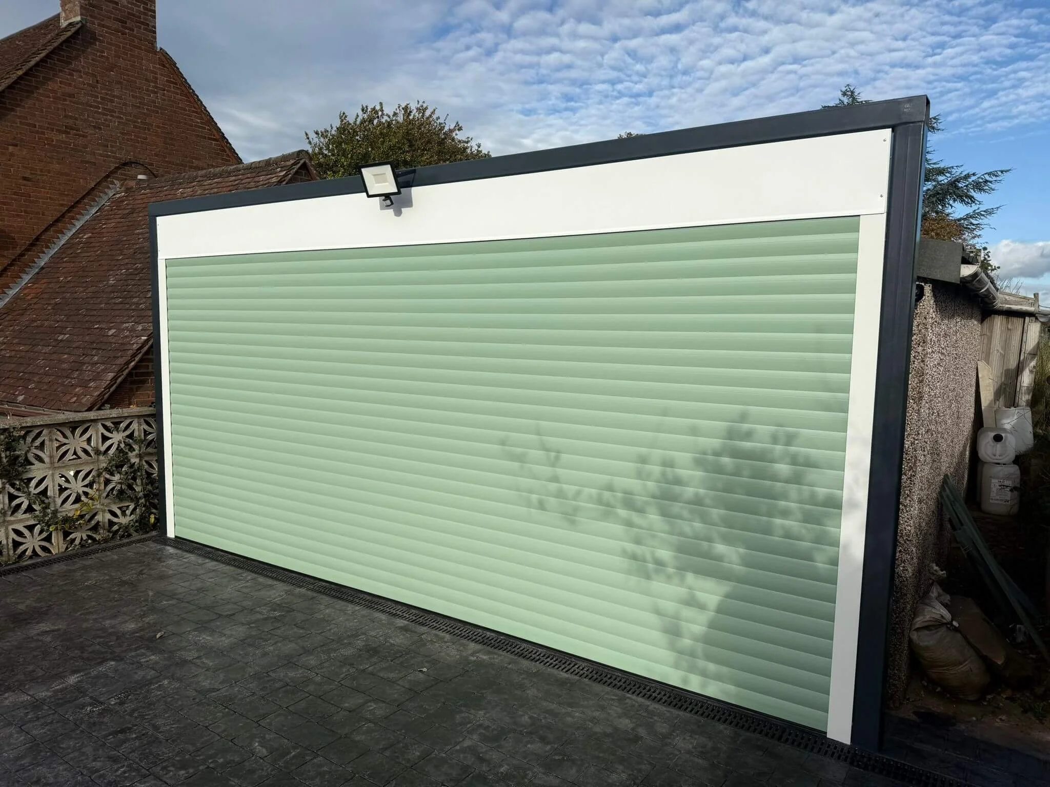 A large, closed green roller shutter on an outdoor building, with a black frame and a white border at the top, under a partly cloudy sky.