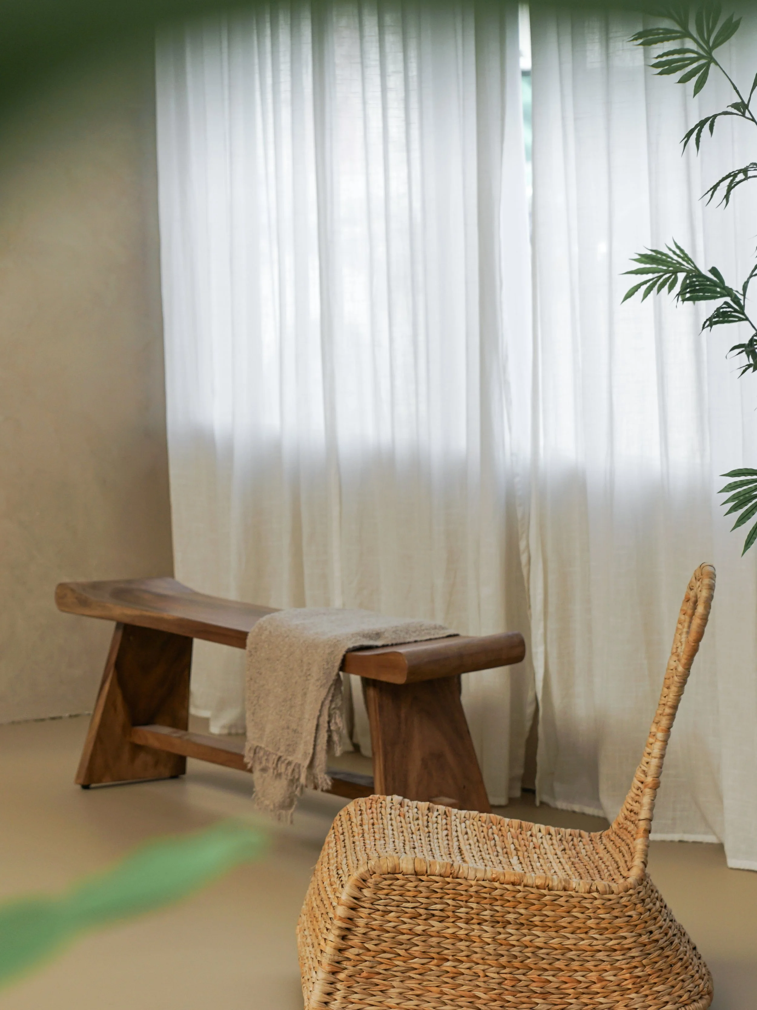 Woonkamer met houten tafel, stoffen deken, rieten stoel en kantoorkruk voor grote ramen met witte gordijnen en binnenplant.