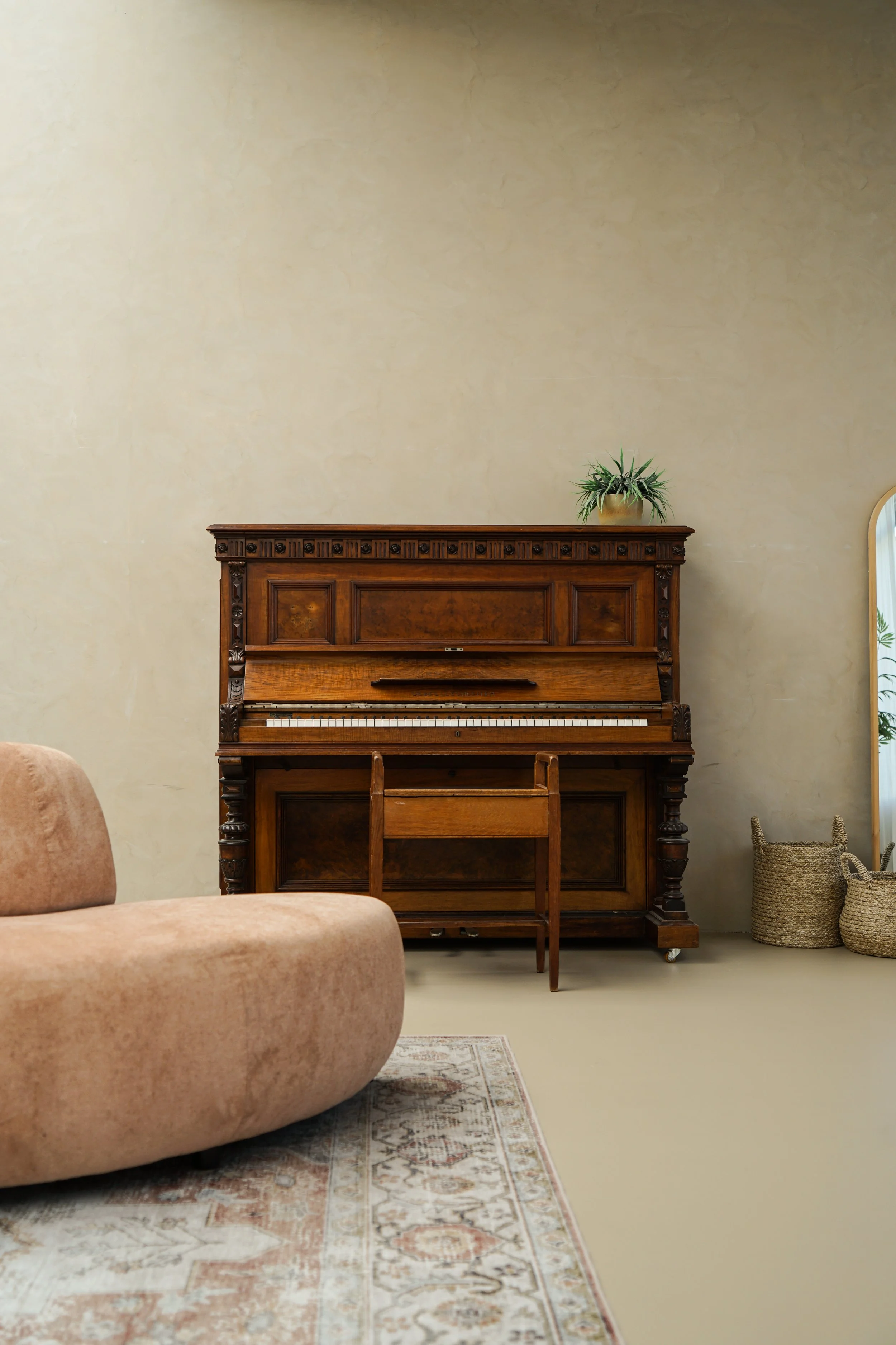 Een vintage houten piano met een groene plant op de bovenkant, een beige bank en een sierlijke stoffen tapijt op de vloer in een woonkamer.
