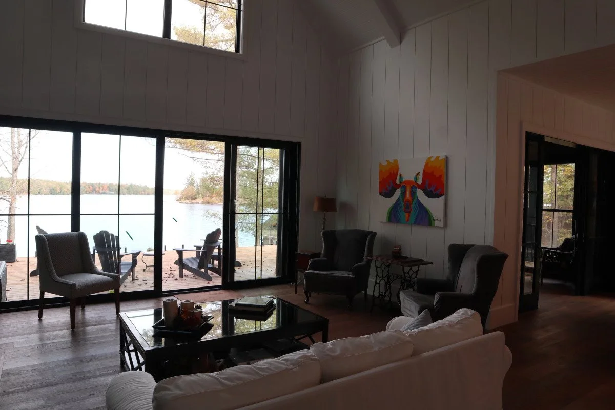 Living room with large glass sliding doors opening to a lake view, with outdoor chairs on a deck. The room has a white sofa, two black armchairs, and a colorful moose painting on the wall.
