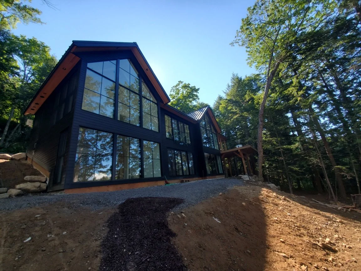 Modern house with large glass windows and a sloped roof, situated on a hillside surrounded by trees under a clear blue sky.