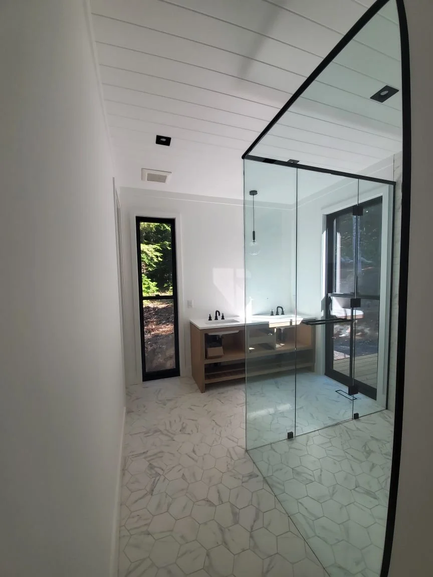 Empty modern bathroom with glass shower enclosure, small white vanity with two sinks, and a window showing greenery outside.