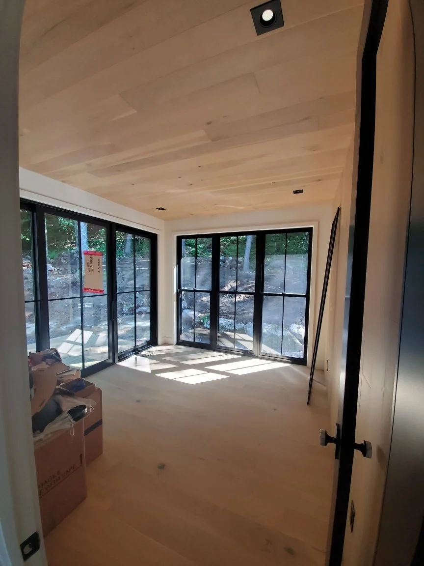 Empty room with wood flooring and ceiling, large black-framed windows showing a rocky outdoor landscape, cardboard boxes in the corner, and construction tools leaning against the wall.