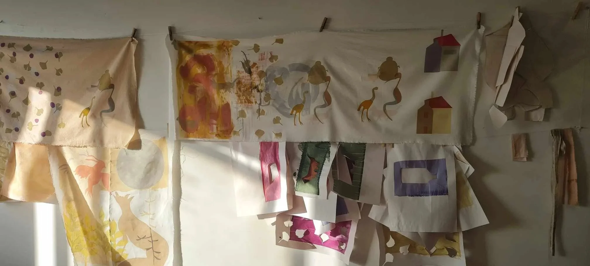 Various pieces of paper with painted and printed designs, depicting animals and houses, hanging on a string with clothespins against a plain wall.