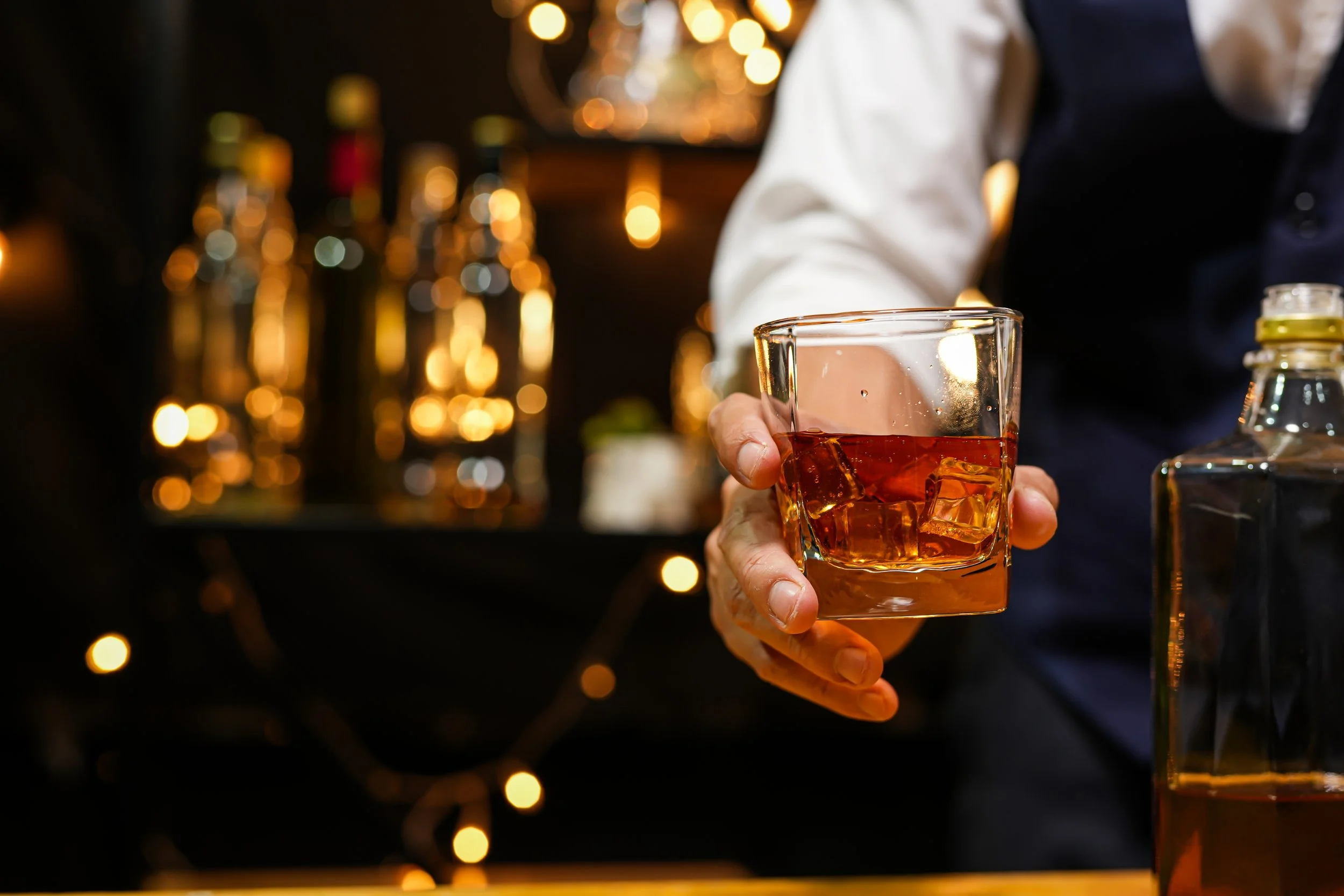 A person in a white shirt and dark vest holding a glass of whiskey on the rocks in a dimly lit bar with blurred warm lights in the background.