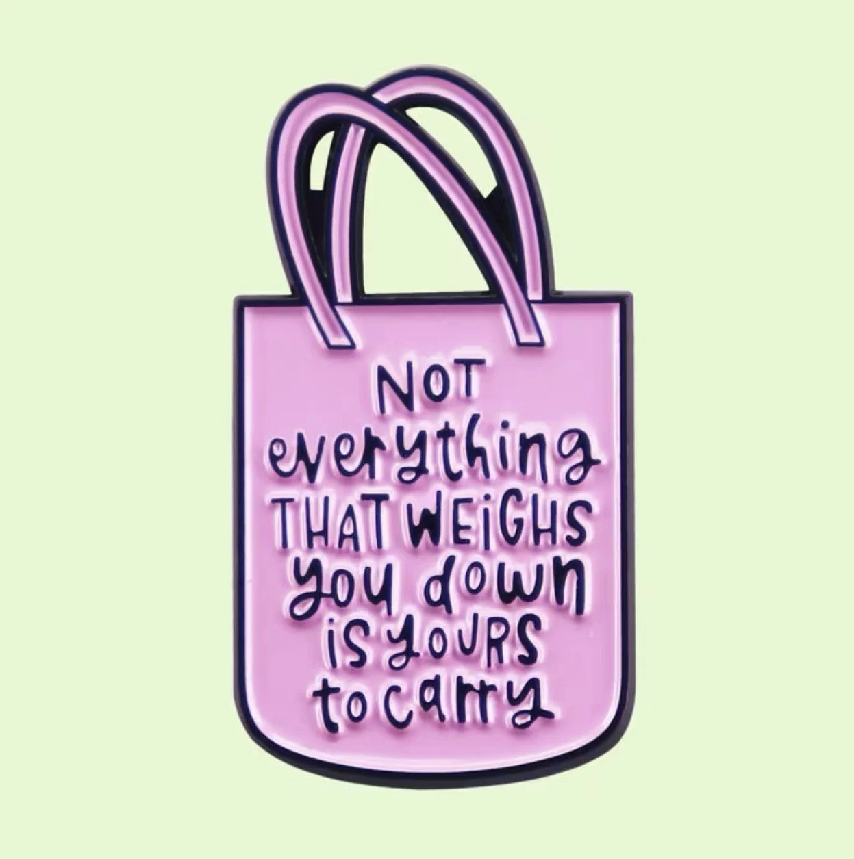 A pin badge in the shape of a pink shopping bag with black outlines. Inside the bag, there is a handwritten message in black that reads: "Not everything that weighs you down is yours to carry." Mental Health (Anchored Thoughts)