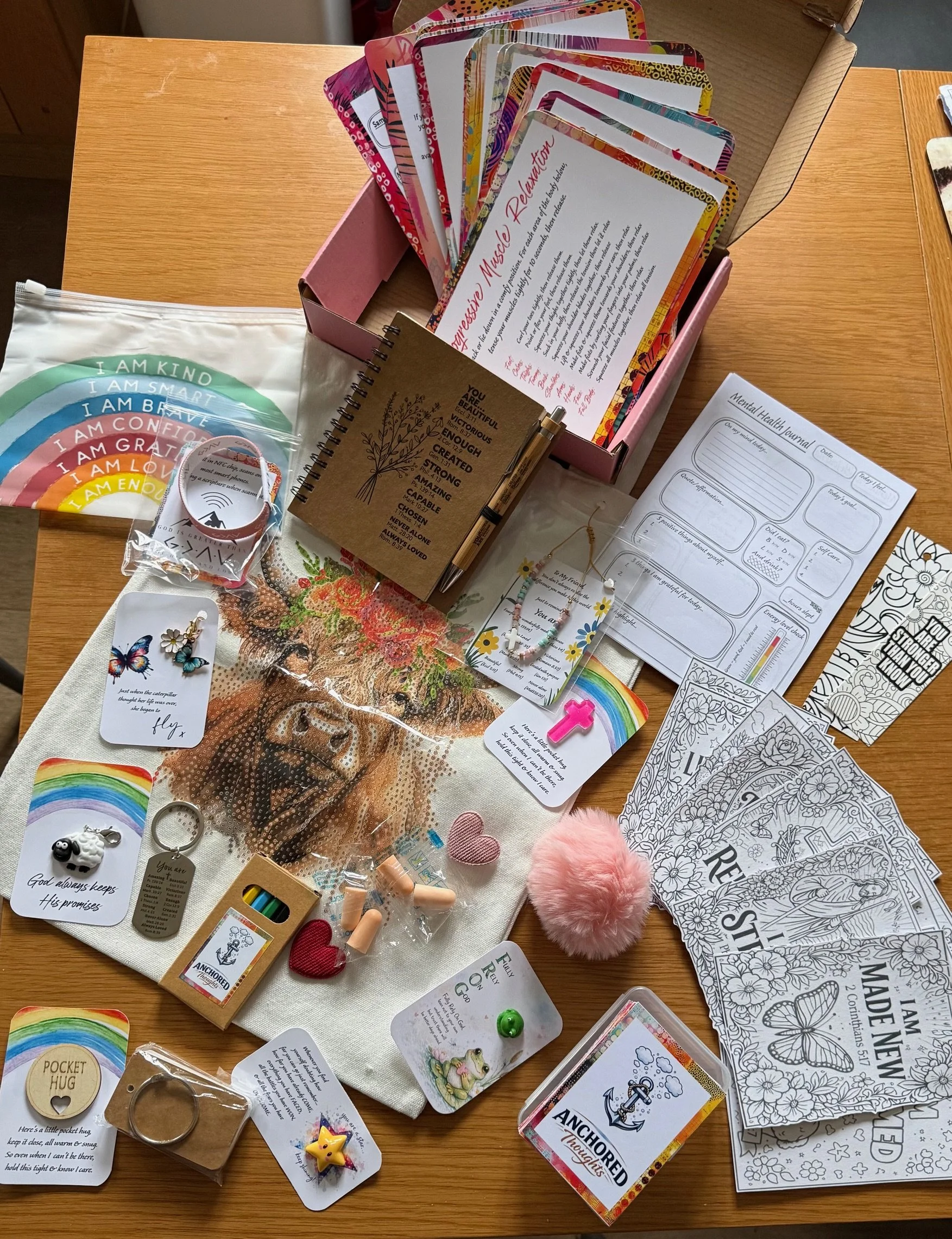 A wooden table with a variety of items including inspirational and motivational cards, coloring pages, decorative jewelry, small toys, and accessories. There are also rainbow stickers, a pink fluffy pom-pom, and a few small trinkets, along with a pink box filled with more cards or organizers.