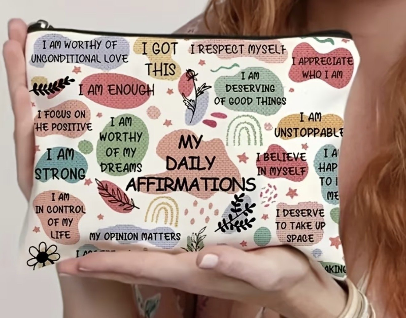 A person holding a colorful affirmations notebook with positive statements about self-worth, strength, appreciation, and dreams, decorated with illustrations of leaves, rainbows, and stars.