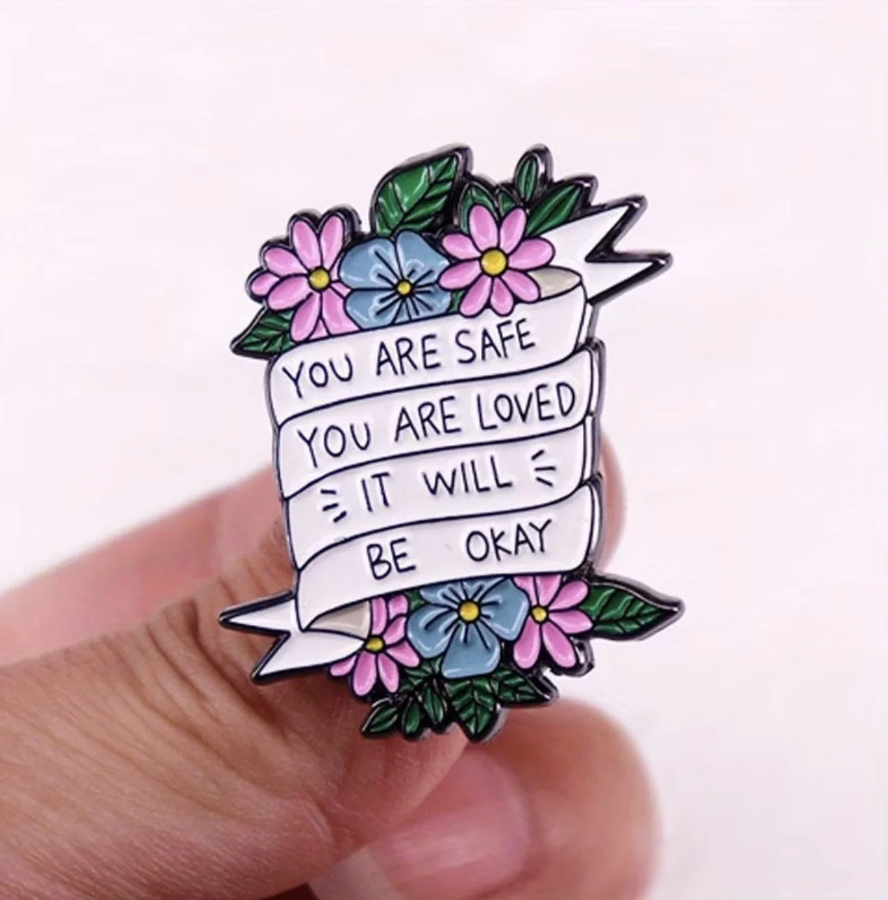 A colourful enamel pin featuring flowers and a banner with the message, "You are safe, you are loved, it will be okay." Mental Health (Anchored Thoughts)