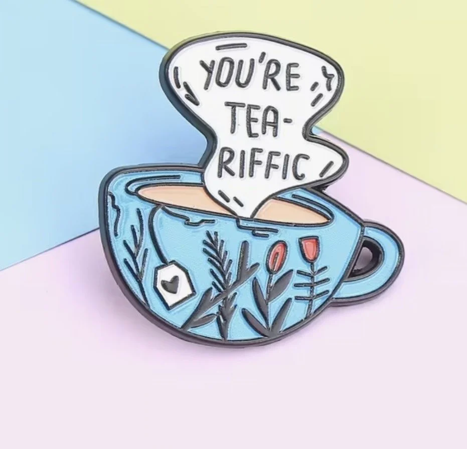 Colorful enamel pin in the shape of a blue teacup with floral and plant designs, showcasing a steam-shaped speech bubble saying 'You're Tea-riffic'. Mental Health (Anchored Thoughts)