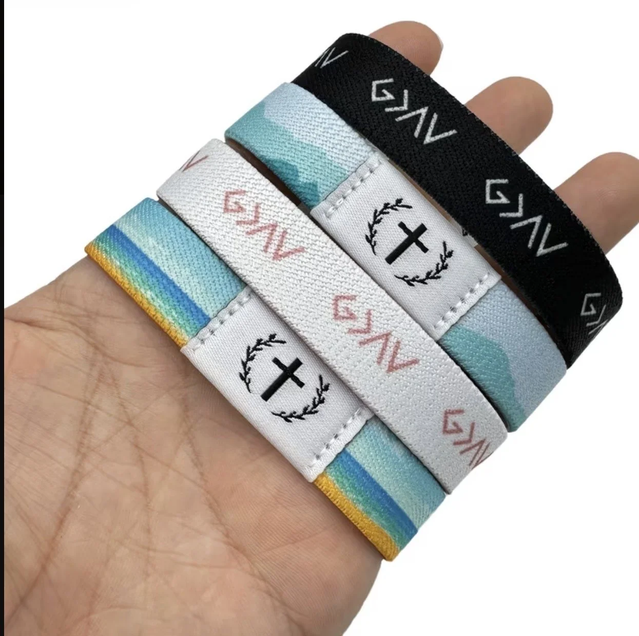 Four fabric wristbands with religious and decorative designs, held in a person's hand. The top wristband is black with white patterns, the second is blue with white and teal, the third is white with a cross and laurel design with red text, and the bo