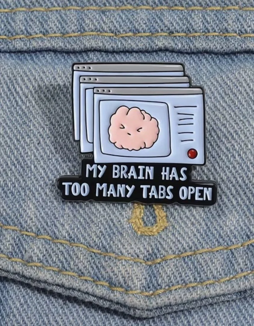 A pin on denim fabric showing a cartoon brain in a windowed monitor with the caption "My brain has too many tabs open".