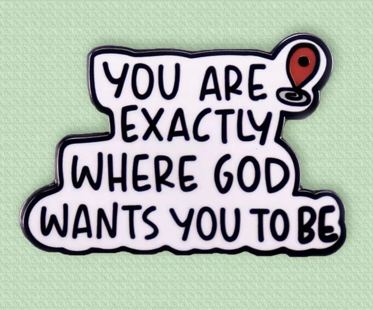 A decorative sign with the words 'You are exactly where God wants you to be' written in black, outlined in black with a stylized red location pin icon at the top right corner, against a light green textured background.