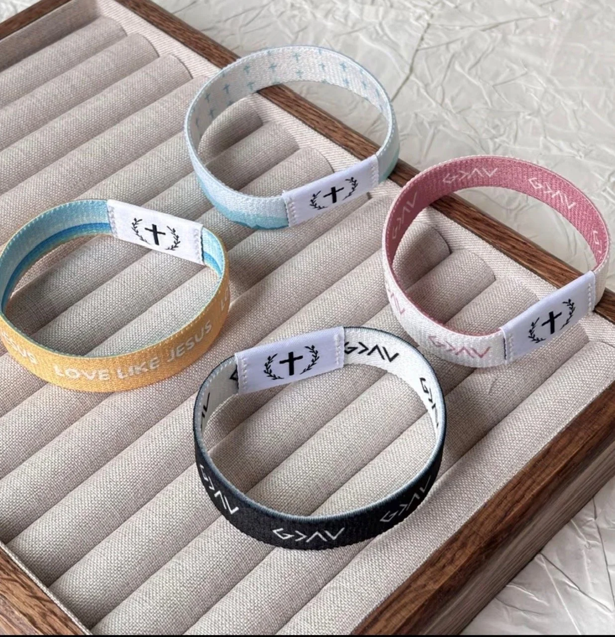 Four fabric headbands with Christian symbols and messages, arranged on a beige cushioned display tray. The headbands are yellow, blue, pink, and black, each with a white fabric patch featuring a black cross surrounded by decorative laurel branches. T