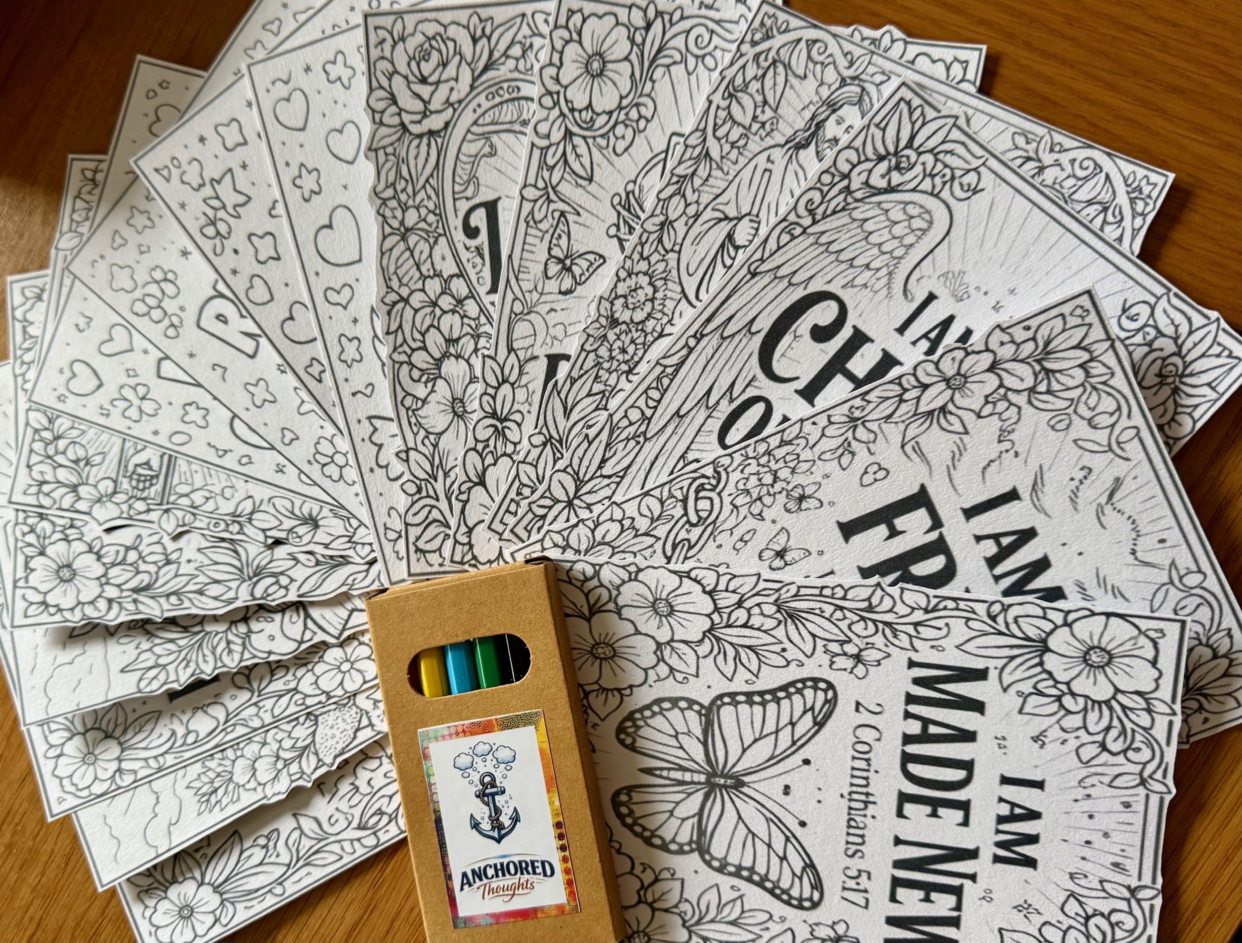 A collection of black and white coloring pages featuring floral and butterfly designs, spread out on a wooden surface. There is also a set of colored markers and a bookmark with an anchor illustration that reads 'Anchored Thoughts'.