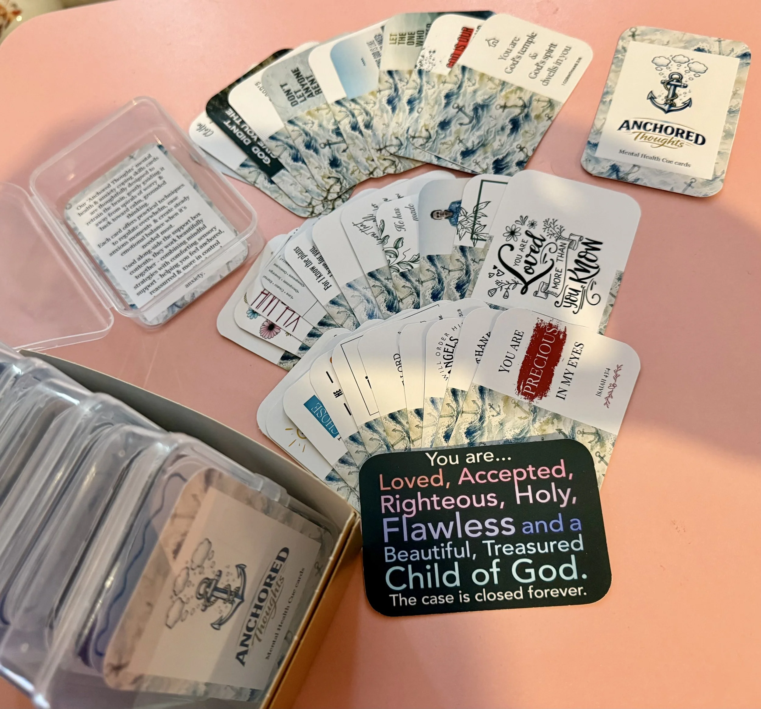 A deck of anchor-themed mental health cue cards fanned out on a pink surface, with a clear plastic container holding additional cards nearby.