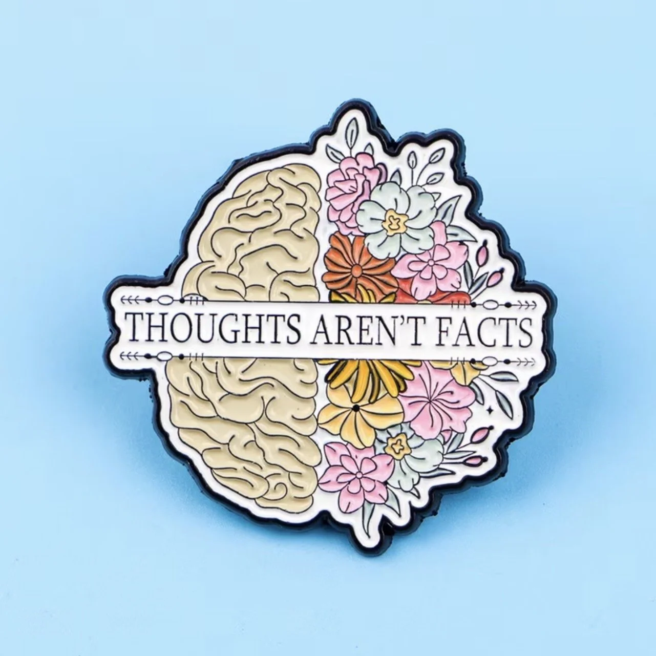 A decorative Mental Health- pin badge  with a brain on the left and colourful flowers on the right, with the phrase "Thoughts Aren't Facts" across the middle. (Anchored Thoughts)