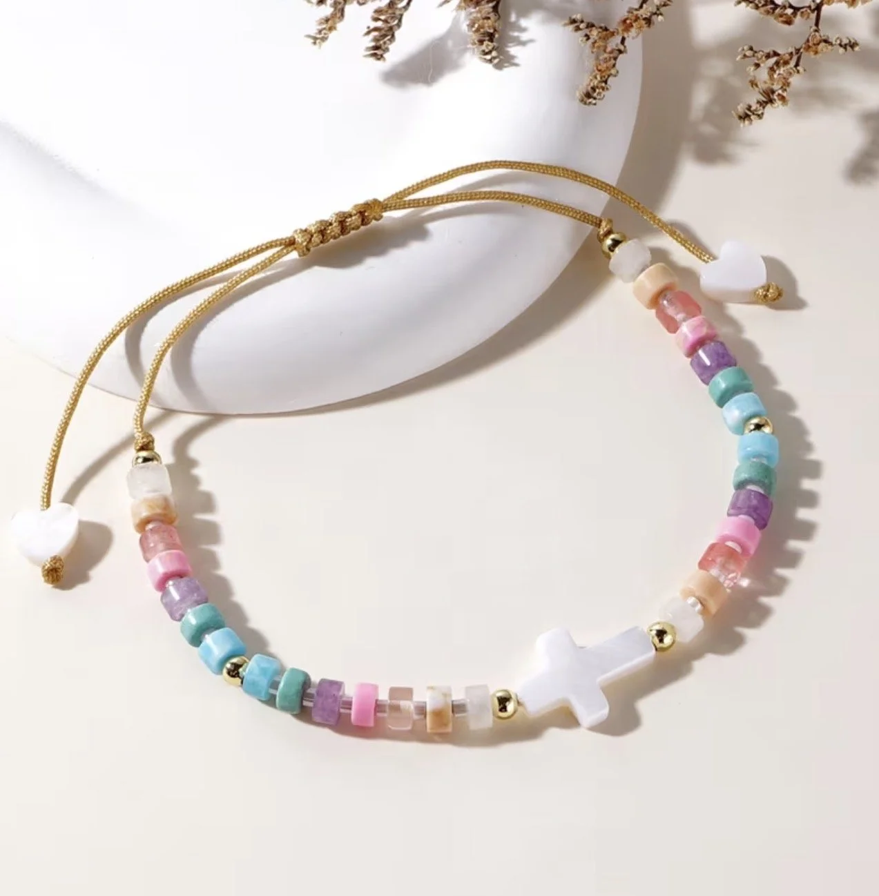 Colorful bead necklace with a white cross pendant and gold accents, displayed on a white surface near a white vase with dried flowers.