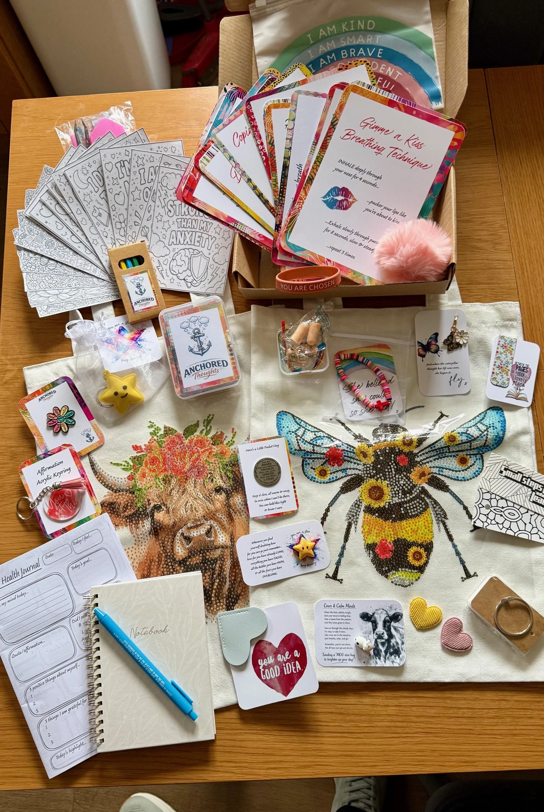 A wooden table with various items, including a partially opened health journal, a blue pen, motivational cards, small plush toys, and a notebook with pages visible. A cloth with an embroidered bee design is also on the table.