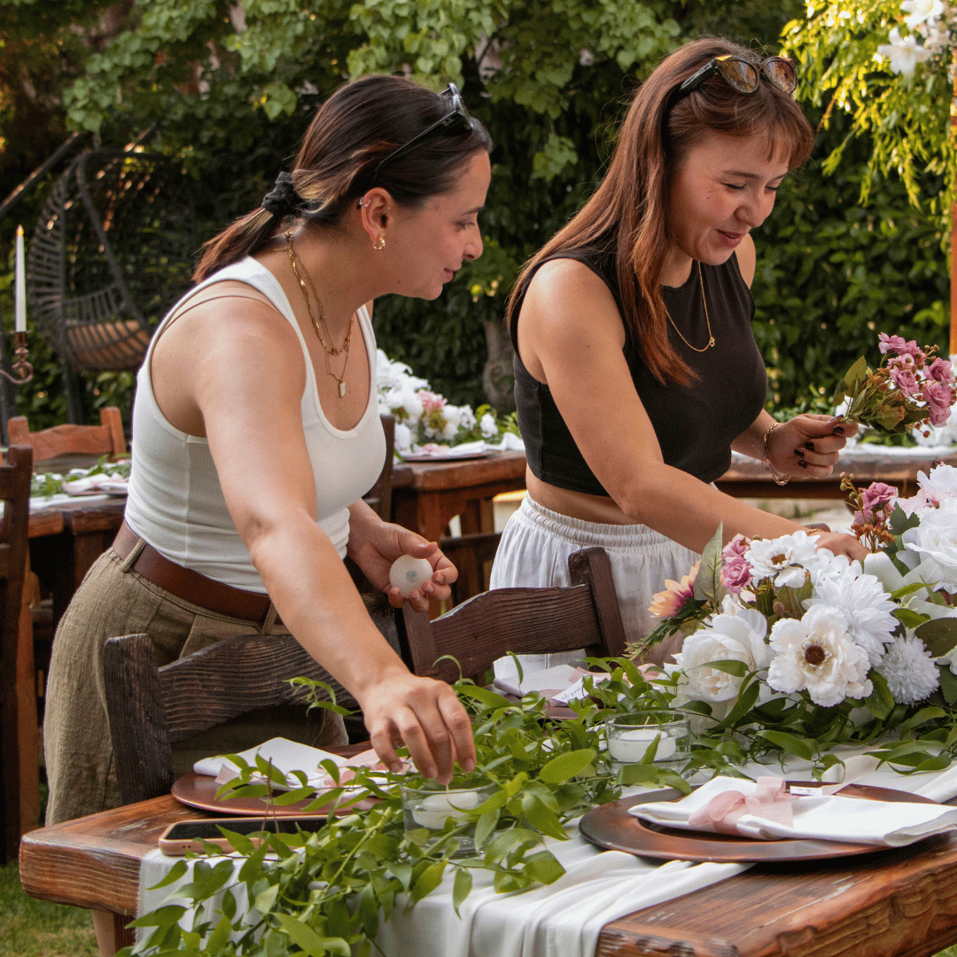 Two people arranging ready-to-use wedding flowers and greenery on an outdoor wooden farm table, representing the local delivery and customer-led setup service from Breemar Flower Farm in Ashland, VA.