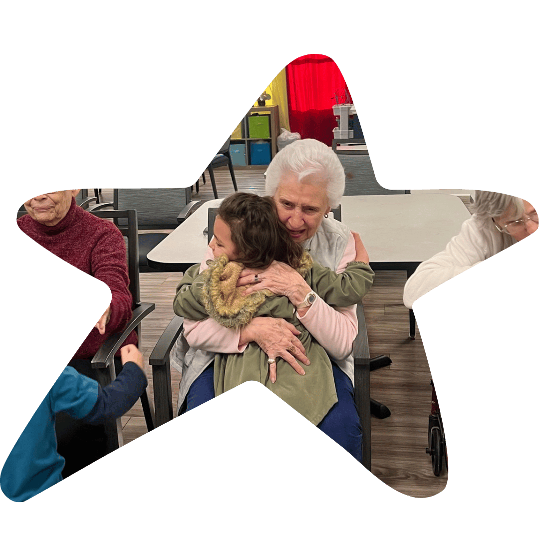 An elderly woman with white hair embracing a woman with dark hair in a hug in a room with other elderly women.