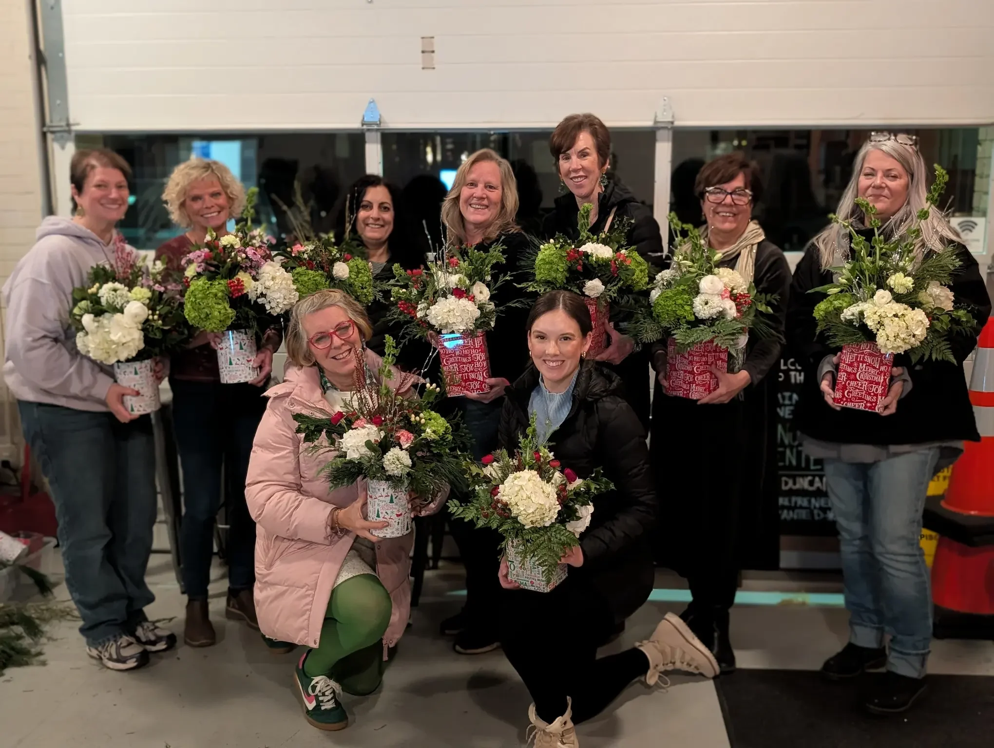 A group of five smiling women participating in a "Petal Party" floral design workshop at Breemar Flower Farm in Ashland, VA, proudly holding their handmade seasonal flower arrangements featuring white hydrangeas, red berries, and evergreens.