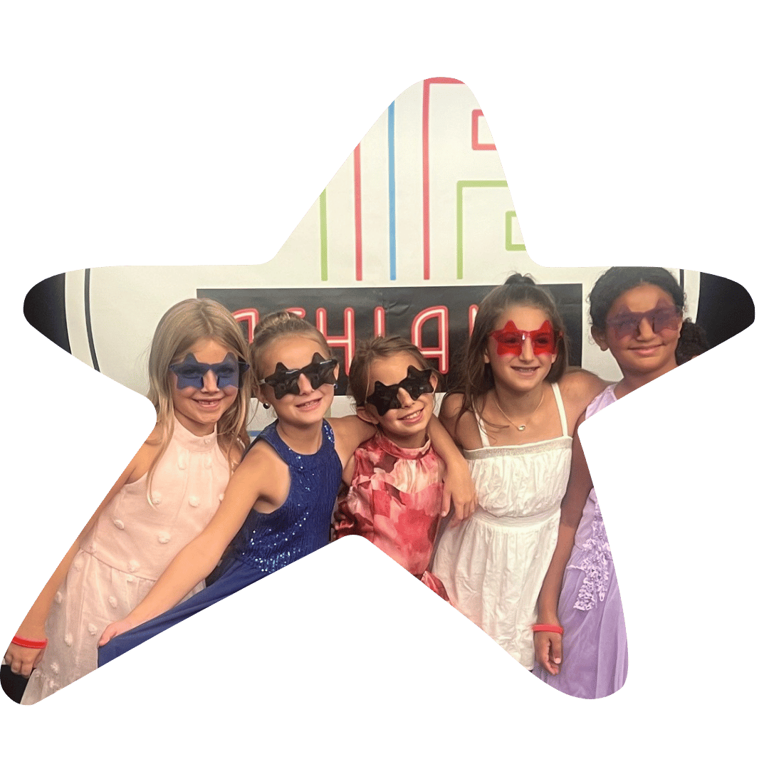 Five young girls wearing colorful star-shaped sunglasses, standing together at a party or event with a colorful wall and a sign in the background.