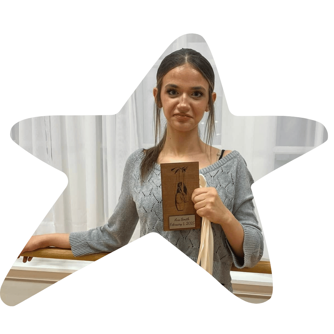 A young woman with long brown hair in a ponytail holding a wooden plaque with an illustration and text that reads "Ana Smith, February 1, 2025". She is wearing a gray sweater and standing in front of white curtains.