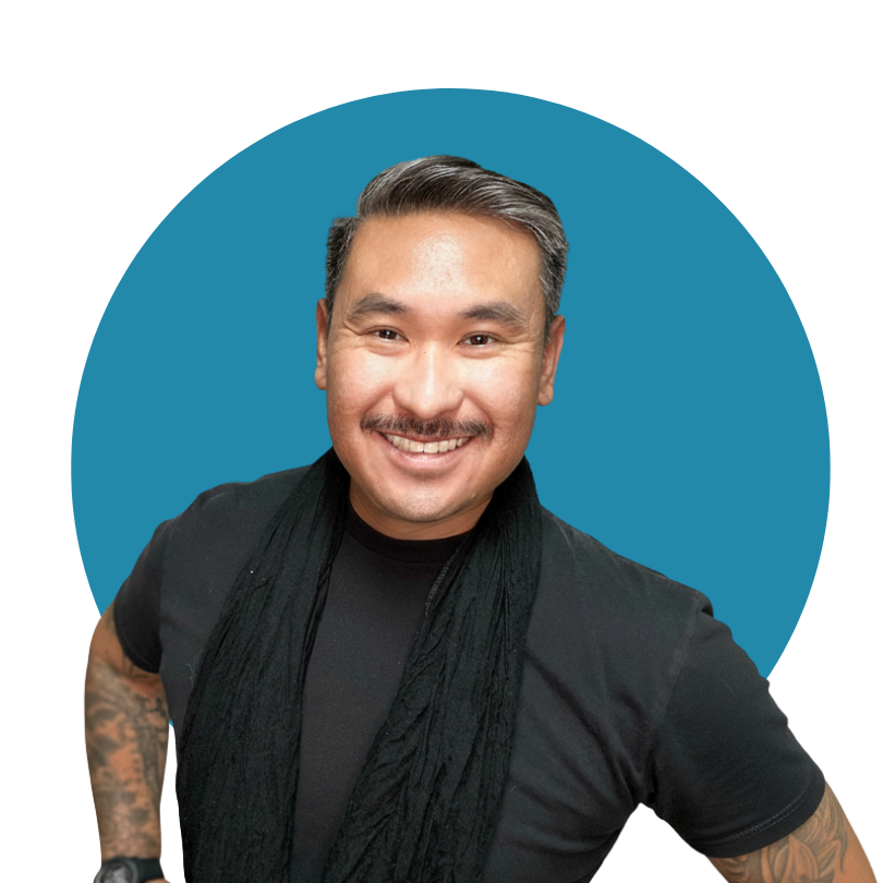 Smiling man with dark hair and tattoos on his left arm, wearing a black shirt and scarf, standing against a blue background.
