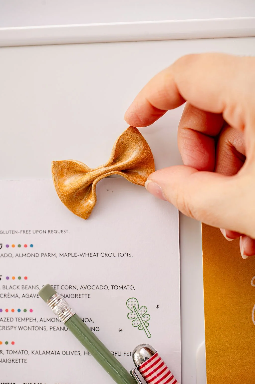 A hand holding a card with a ceramic pasta fridge magnet attached. The card has text that reads 'CERAMIC PASTA FRIDGE MAGNET glazed stoneware' and an illustration of a wine glass, with a small fork and knife icons at the bottom.