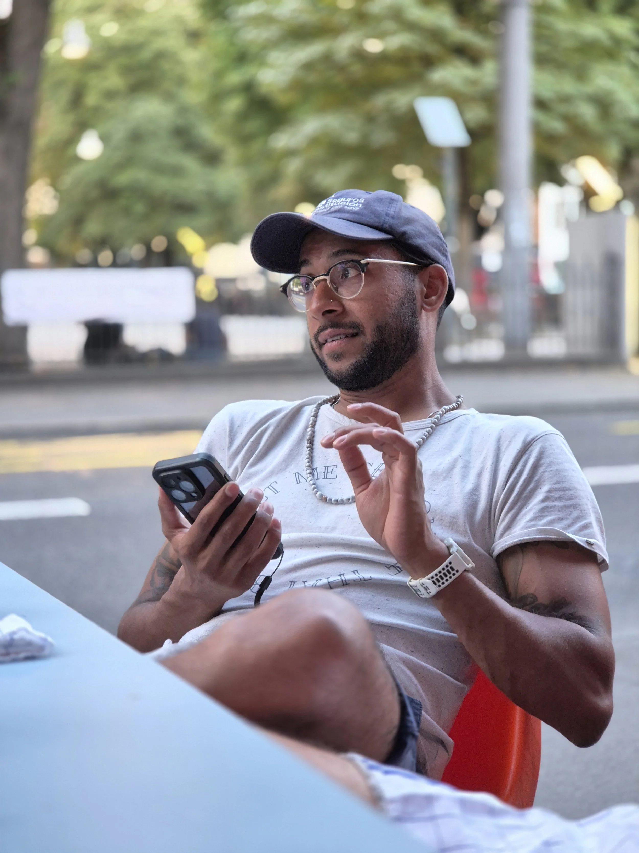 Ein junger Mann mit Brille, Bart, dunkelblauer Baseballkappe und T-Shirt sitzt draußen am Tisch, hält ein Smartphone in der Hand, schaut überrascht oder verwirrt und gestikuliert mit der anderen Hand. Im Hintergrund sind Bäume, eine Straße und eine Bushaltestelle sichtbar.
