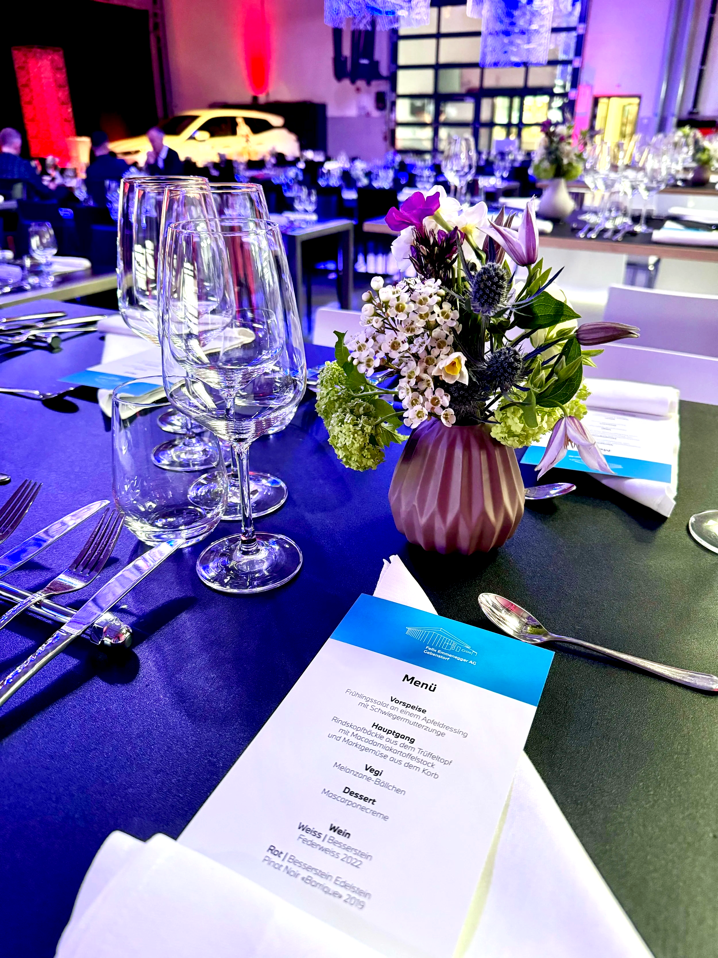 Ein gedeckter Tisch bei einer Veranstaltung mit Gläsern, Besteck, Blumenstrauß in einer Vase und einem Menü in deutscher Sprache, im Hintergrund ein Auto und eine Bühne mit bunten Lichtern.